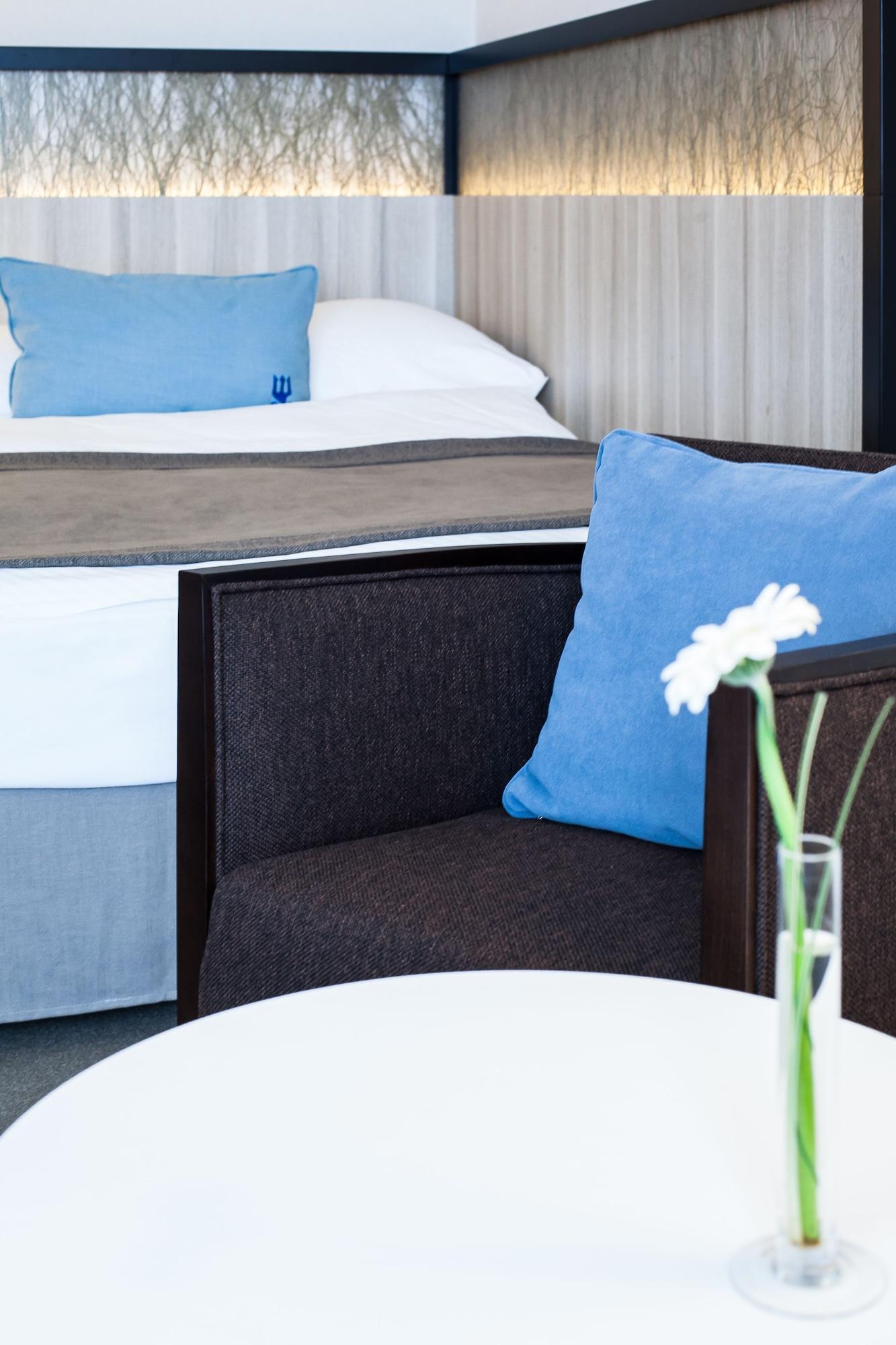 Hotel Neptun Росток Экстерьер фото The photo features a modern hotel room setting. In the foreground, there is a dark-colored chair with a light blue cushion. A small white table is in front of the chair, holding a decorative vase with a single flower. In the background, a neatly made