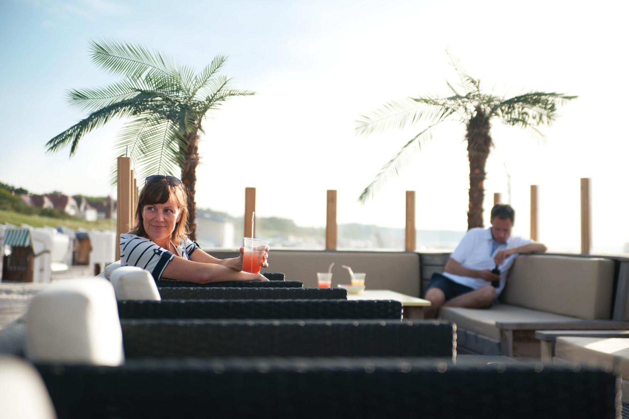 Hotel Neptun Росток Экстерьер фото The photo shows a relaxed outdoor setting, likely on a terrace or patio. In the foreground, a woman with dark hair is sitting at a table, holding a colorful drink and looking directly at the camera. She appears to be enjoying her time. In the backgro